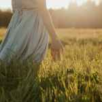 Woman standing in a sunlit meadow with eyes closed and gentle smile, embodying a pet caregiver in the quiet, ongoing stages of grief beginning to feel quietly proud of the love she gave.